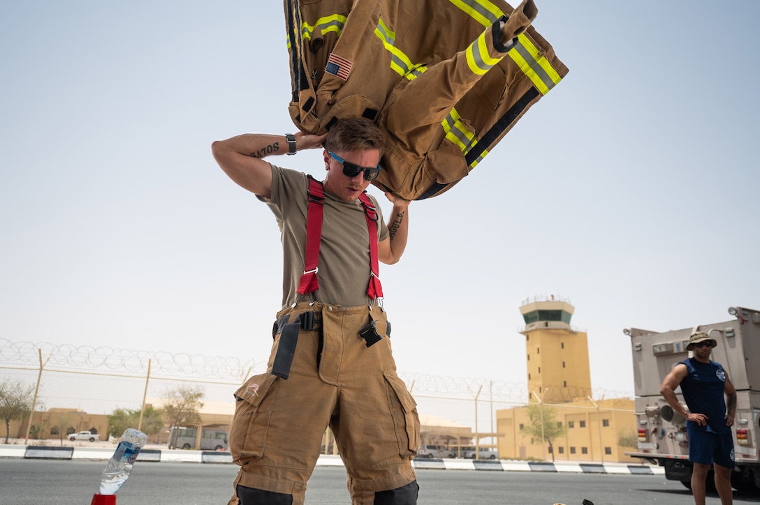 An airman puts on a firefighting jacket in a large parking area.