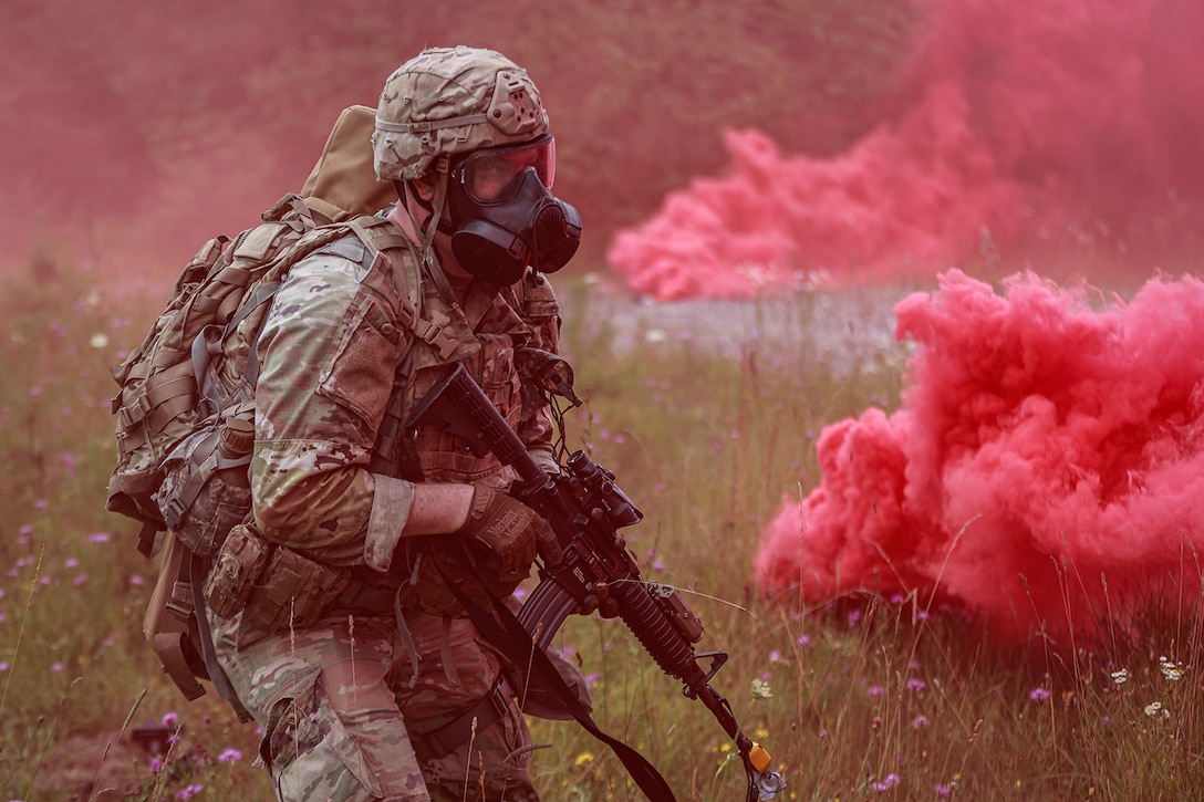 A uniformed soldier wearing a helmet and gas mask walks with a weapon through pink simulated tear gas.