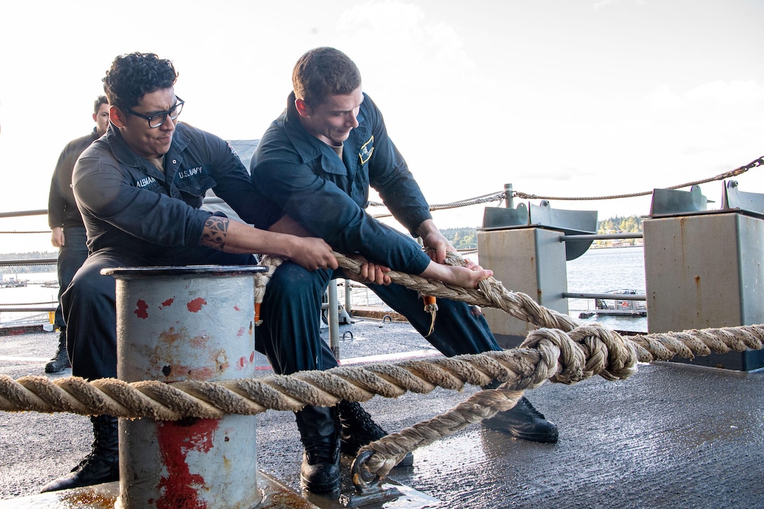 Two sailors lean to the left while pulling a rope aboard a ship as a fellow sailor watches in the background.