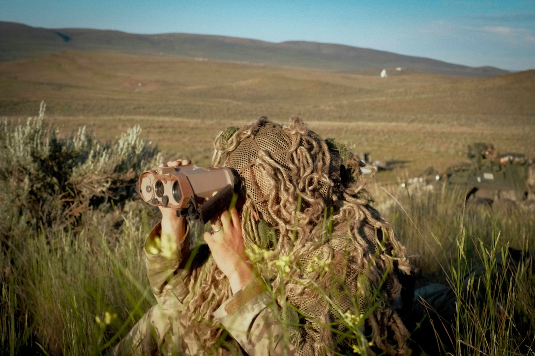 A soldier in camouflage gear uses binoculars while sitting in tall grass in a field with mountains in the background.