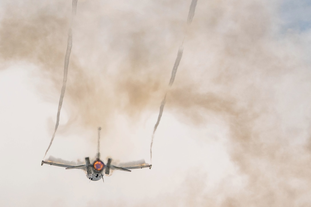 An aircraft leaves streaks while flying through clouds of smoke illuminated by orange rear-engine flames.