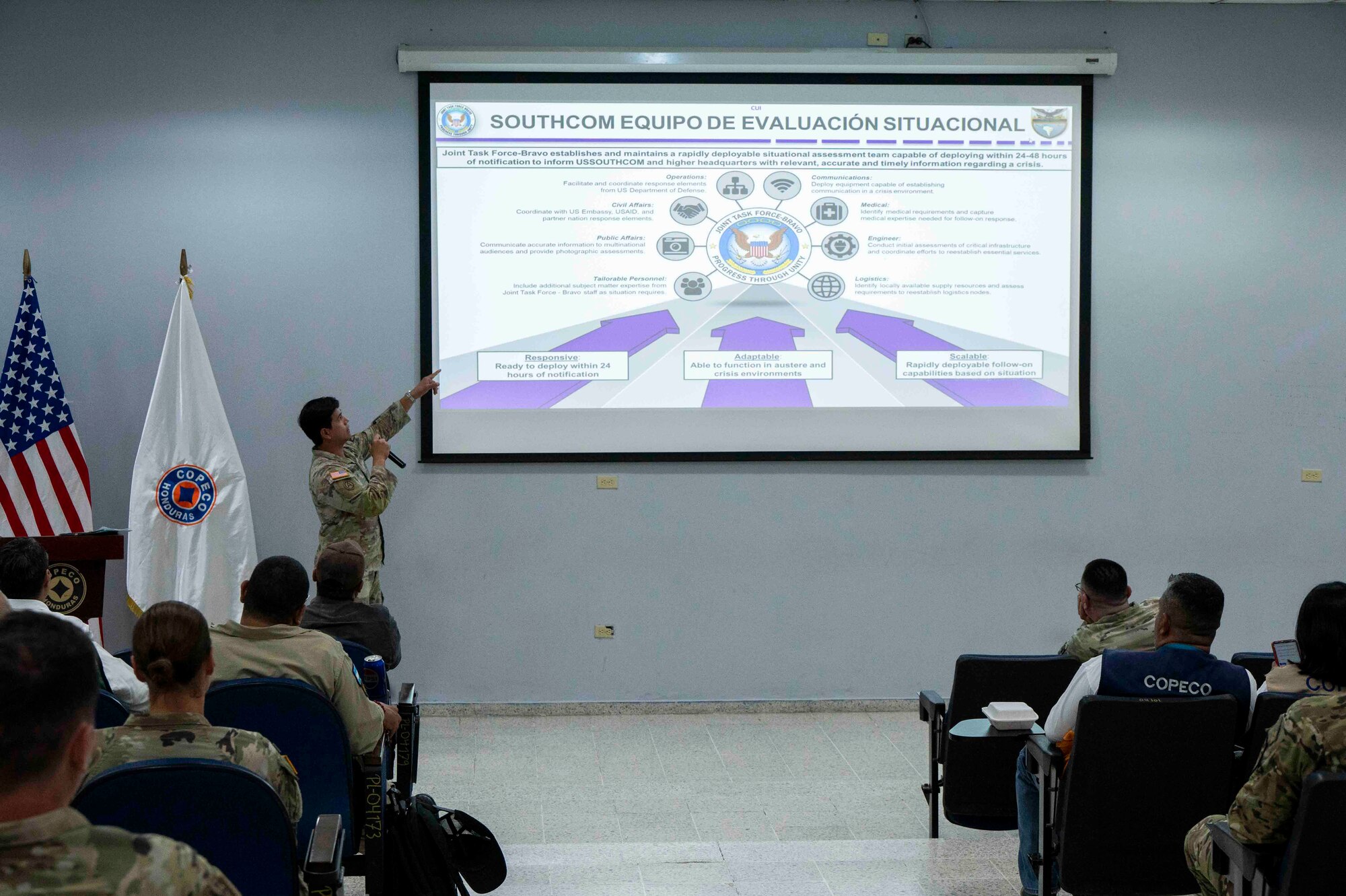 A photo of a military member pointing at a slide during a presentation.