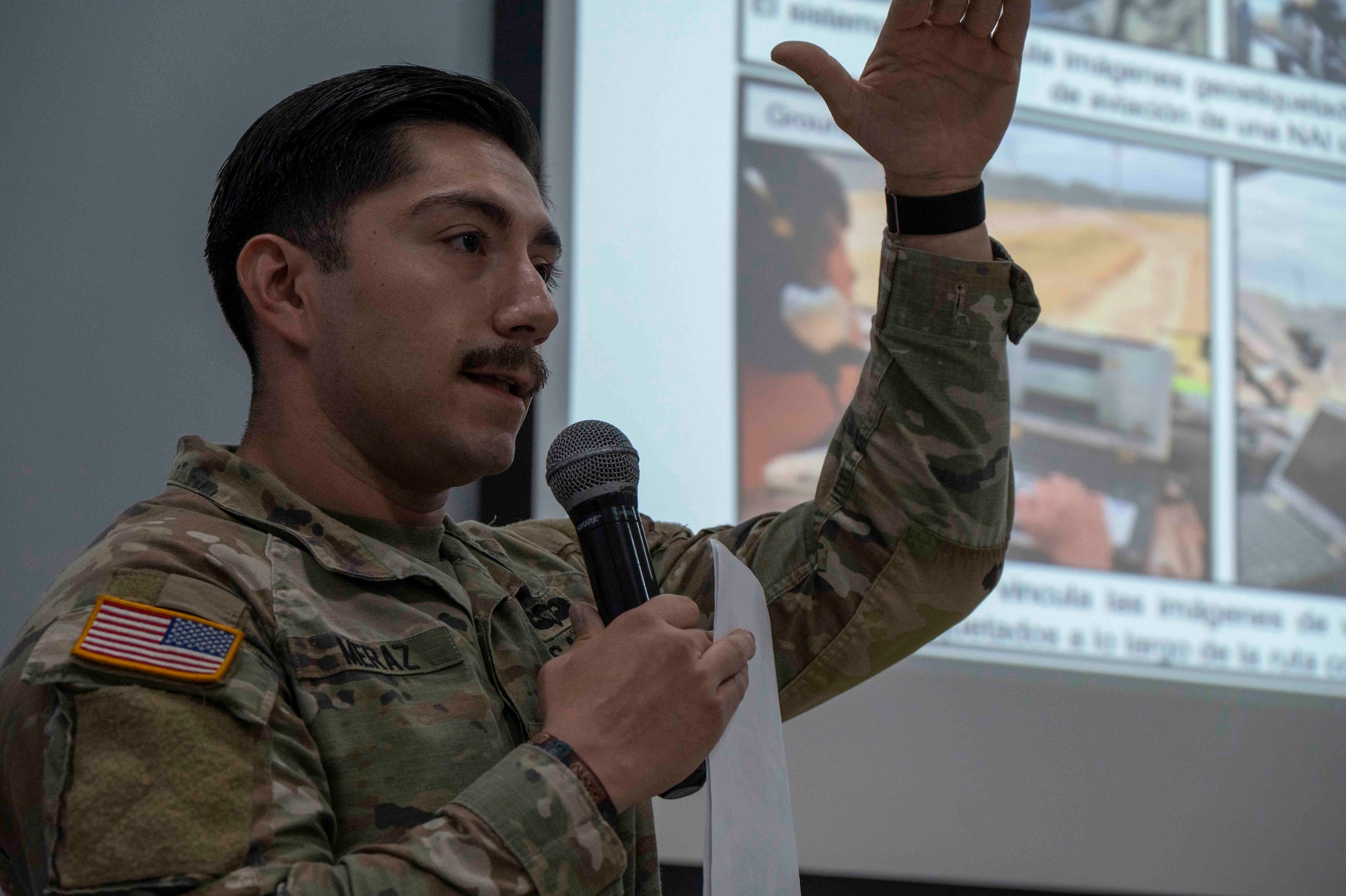 A photo of a military member presenting a slideshow, talking with a microphone and paper in hand.