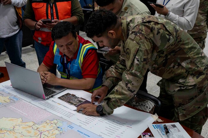 A photo of a military member pointing at a map on a table to another person.
