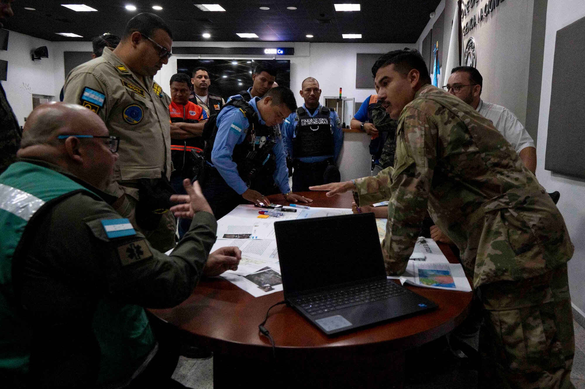 A photo of a military member talking to a group of people around a table with maps and computers.