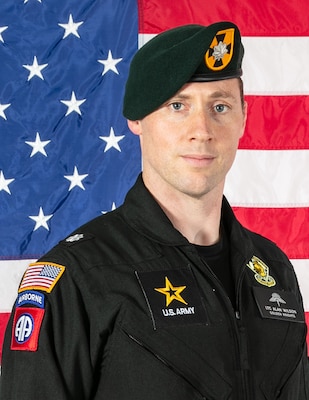man standing in front of two flags in U.S. Army flight suit uniform.