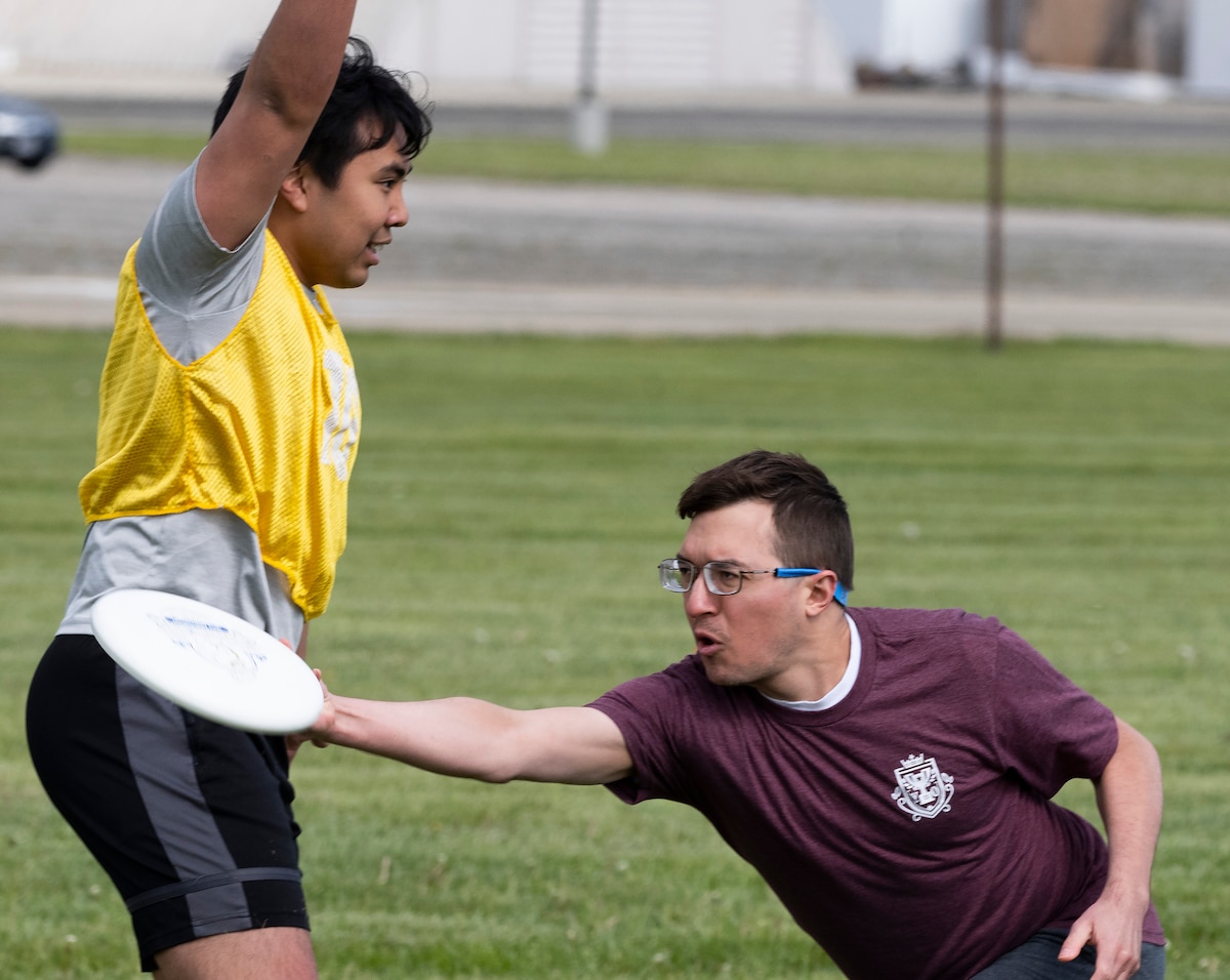 A man in a maroon tee shirt throws a flying disk past a man in a yellow vest who has his arms extended.