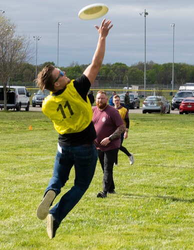 A man in a yellow vest leaps into the air reaching for a flying disk.