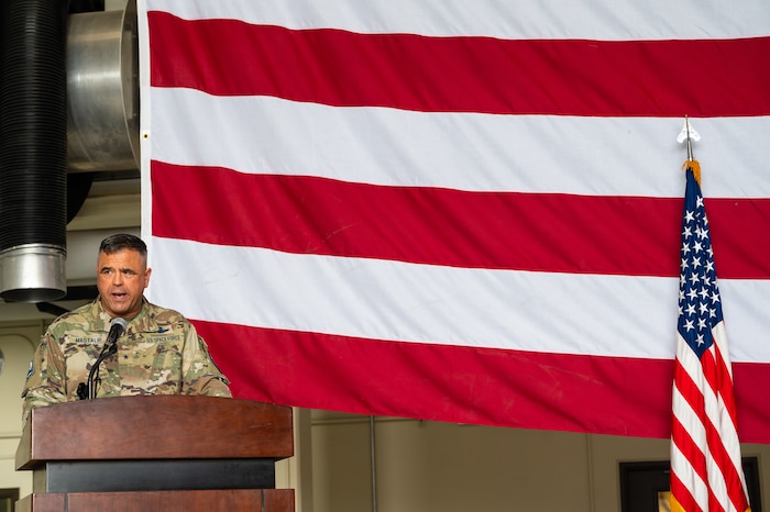 A uniformed man in camouflage stands behind a wooden podium in left corner of a photo, with a large U.S. flag hanging horizontally behind him and the U.S. flag hanging on a small post upright on the right side of the photo.