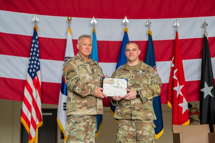 Two men in camouflage uniforms stand together displaying a certificate between them in front of a row of military flags, with a large U.S. flag hanging horizontally behind them as a backdrop.