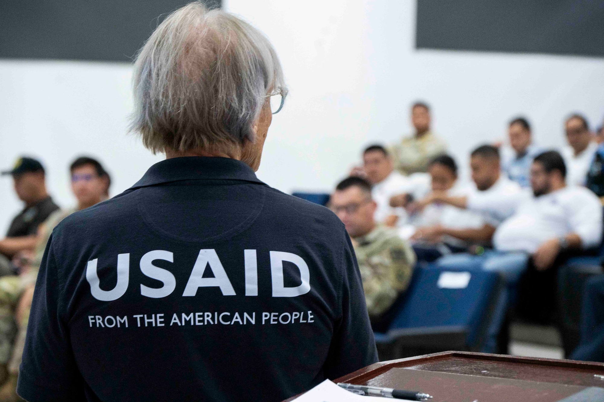 A photo of a person standing facing a crown with "USAID FROM THE AMERICAN PEOPLE" printed on the back of her shirt.