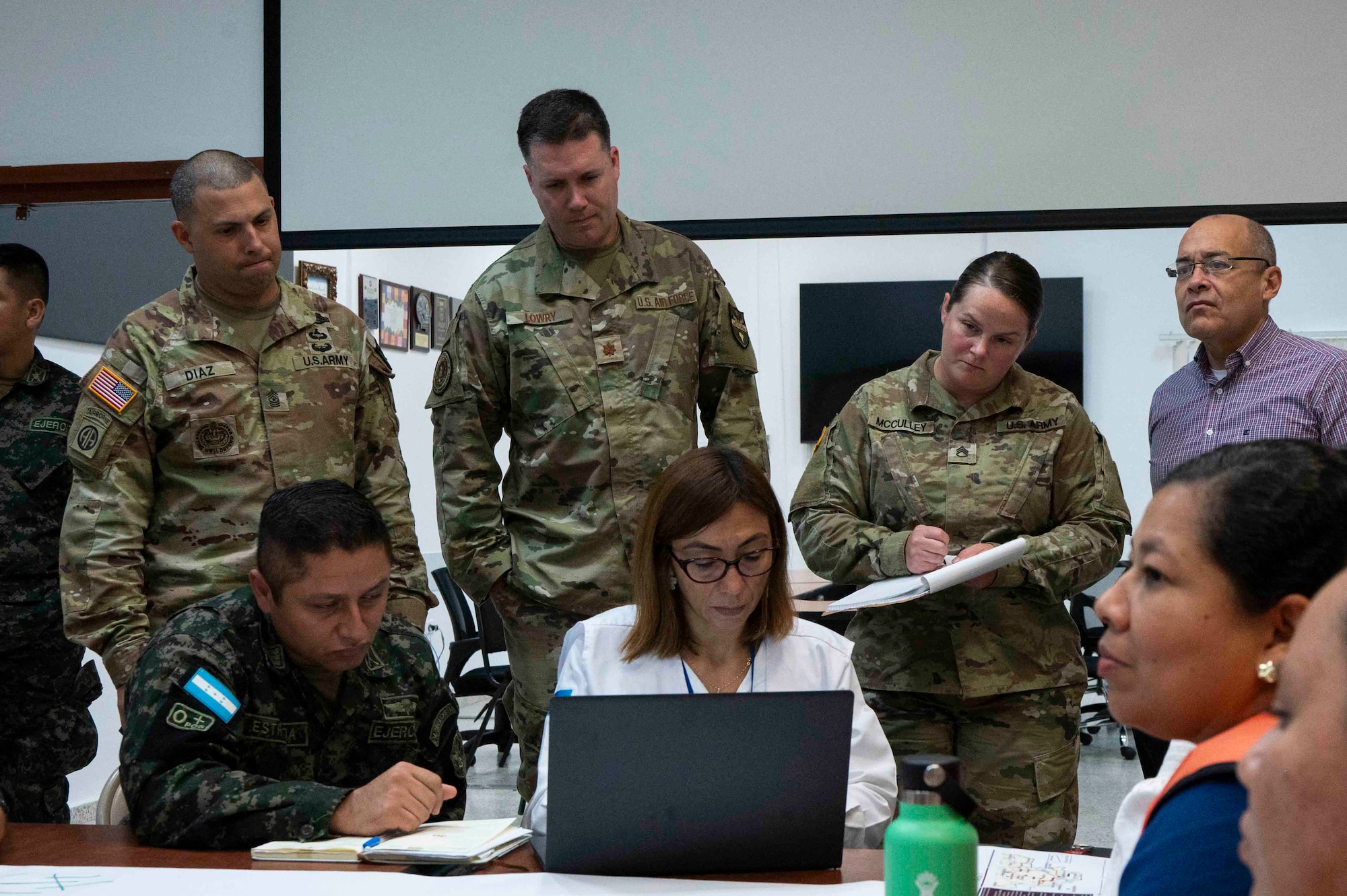 A photo of three military members looking over the shoulder of people on laptops and taking notes.