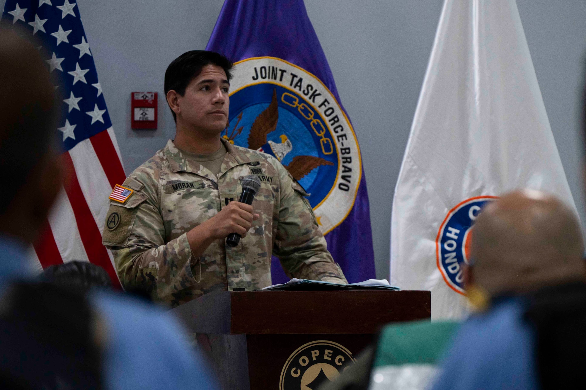 A photo of a military member standing at a podium with a microphone.