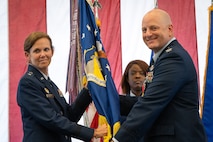 Major General Regina Sabric and Colonel Jason grandy are both in full service dress while holding a military guideon. A chief is standing behind them and a large American flag is draped in the background.