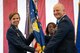 Major General Regina Sabric and Colonel Jason grandy are both in full service dress while holding a military guideon. A chief is standing behind them and a large American flag is draped in the background.