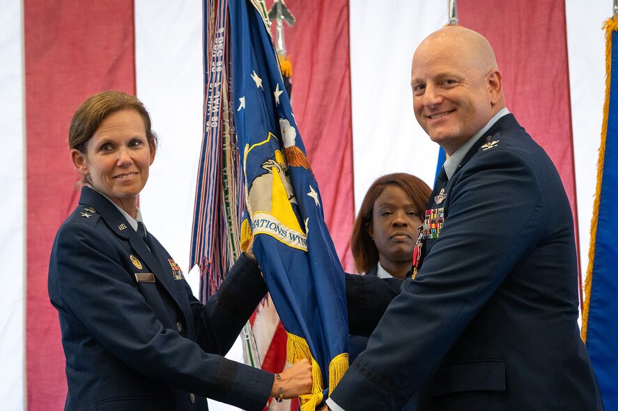Major General Regina Sabric and Colonel Jason grandy are both in full service dress while holding a military guideon. A chief is standing behind them and a large American flag is draped in the background.