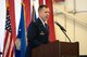 Colonel Scott Hurrelbrink leans on a podium while in full service dress. He is speaking into a microphone while in a large hangar. To his rear are a row of three flags juxtaposed against a large hanging American flag.