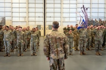 A formation of Airmen render a salute, one of them quickly pushing forward a guideon, in a hangar while overlooked by a colonel.