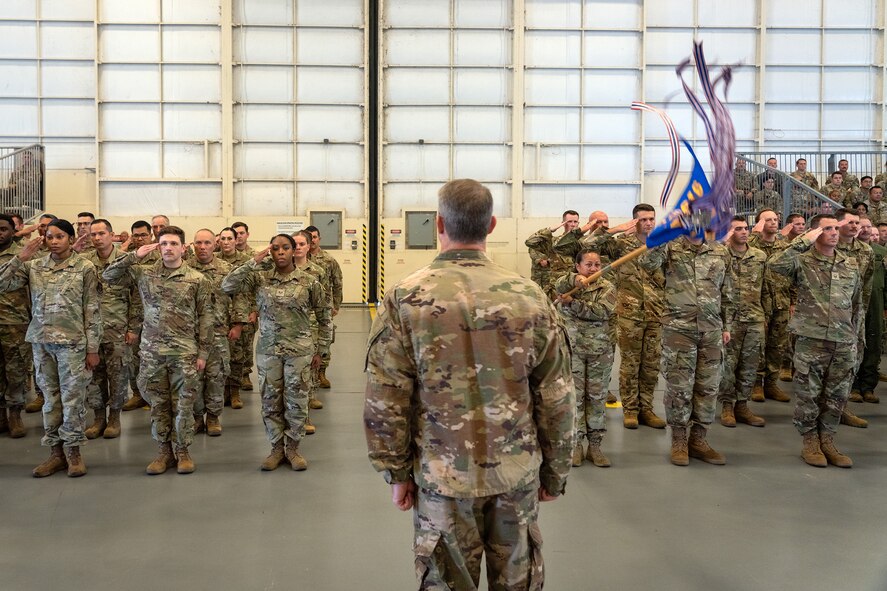 A formation of Airmen render a salute, one of them quickly pushing forward a guideon, in a hangar while overlooked by a colonel.