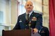 Colonel Jason grandy leans on a podium in his full service dress in a hangar while speaking to the unit during his change of command.
