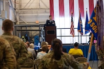 A crowd of Airmen and local civic leaders watch as Colonel Scott Hurrelbrink, in his full service dress, speaks at a on-stage podium in the distance