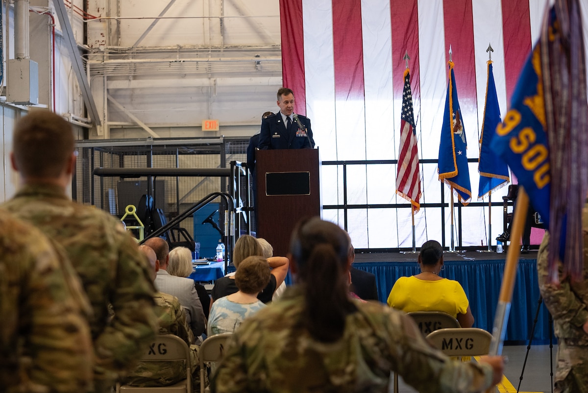 A crowd of Airmen and local civic leaders watch as Colonel Scott Hurrelbrink, in his full service dress, speaks at a on-stage podium in the distance