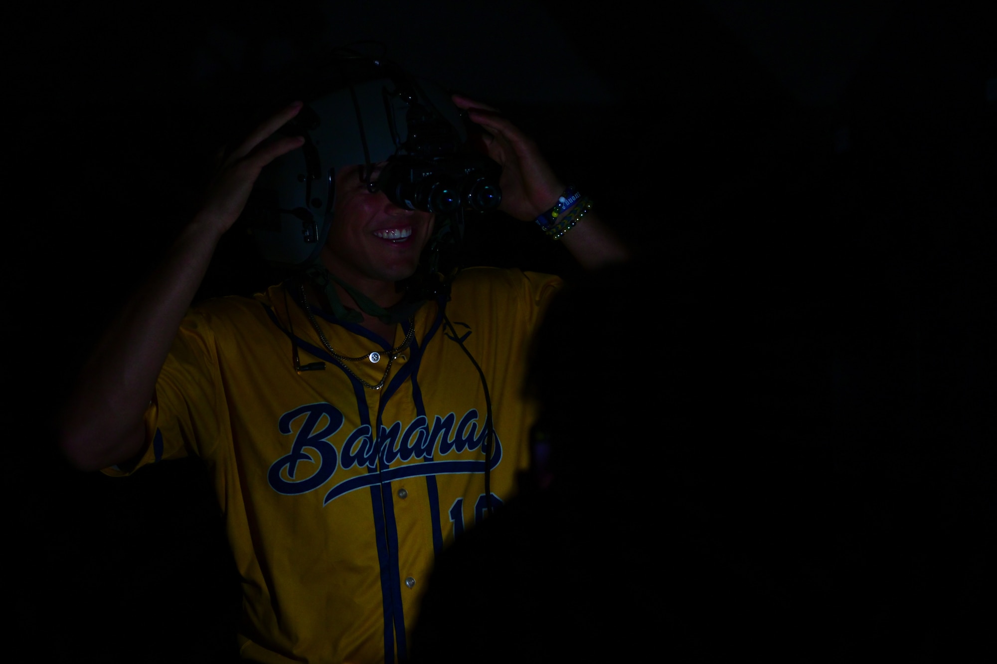 A person wearing a yellow "Savannah Bananas" baseball jersey is adjusting night vision goggles in a dark environment. Only their face and hands are partially illuminated.
