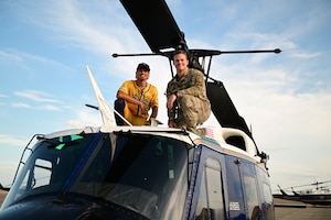 Two individuals, one in a yellow "Savannah Bananas" baseball jersey and the other in military camouflage, are sitting on top of a helicopter. They are both smiling and posing for the camera.