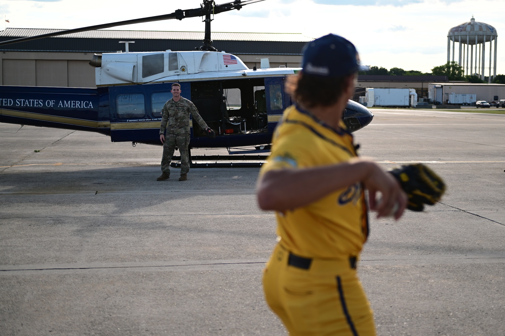 A man in a yellow "Savannah Bananas" baseball jersey is preparing to throw a baseball at a man in military camouflage standing in front of a helicopter.