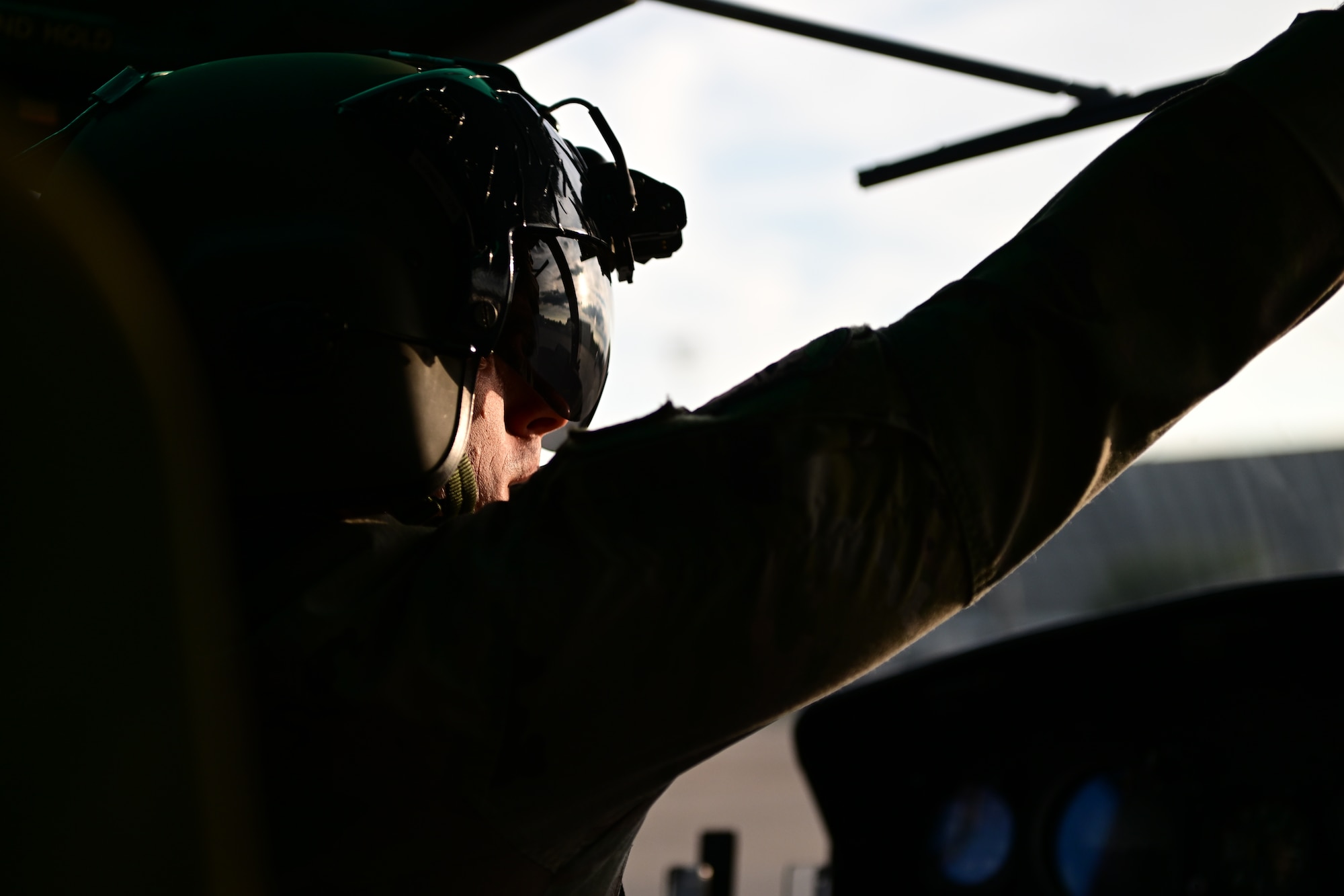 A close-up of a pilot wearing a helmet, with one arm raised, inside a helicopter cockpit. The sunlight casts a shadow over their face.