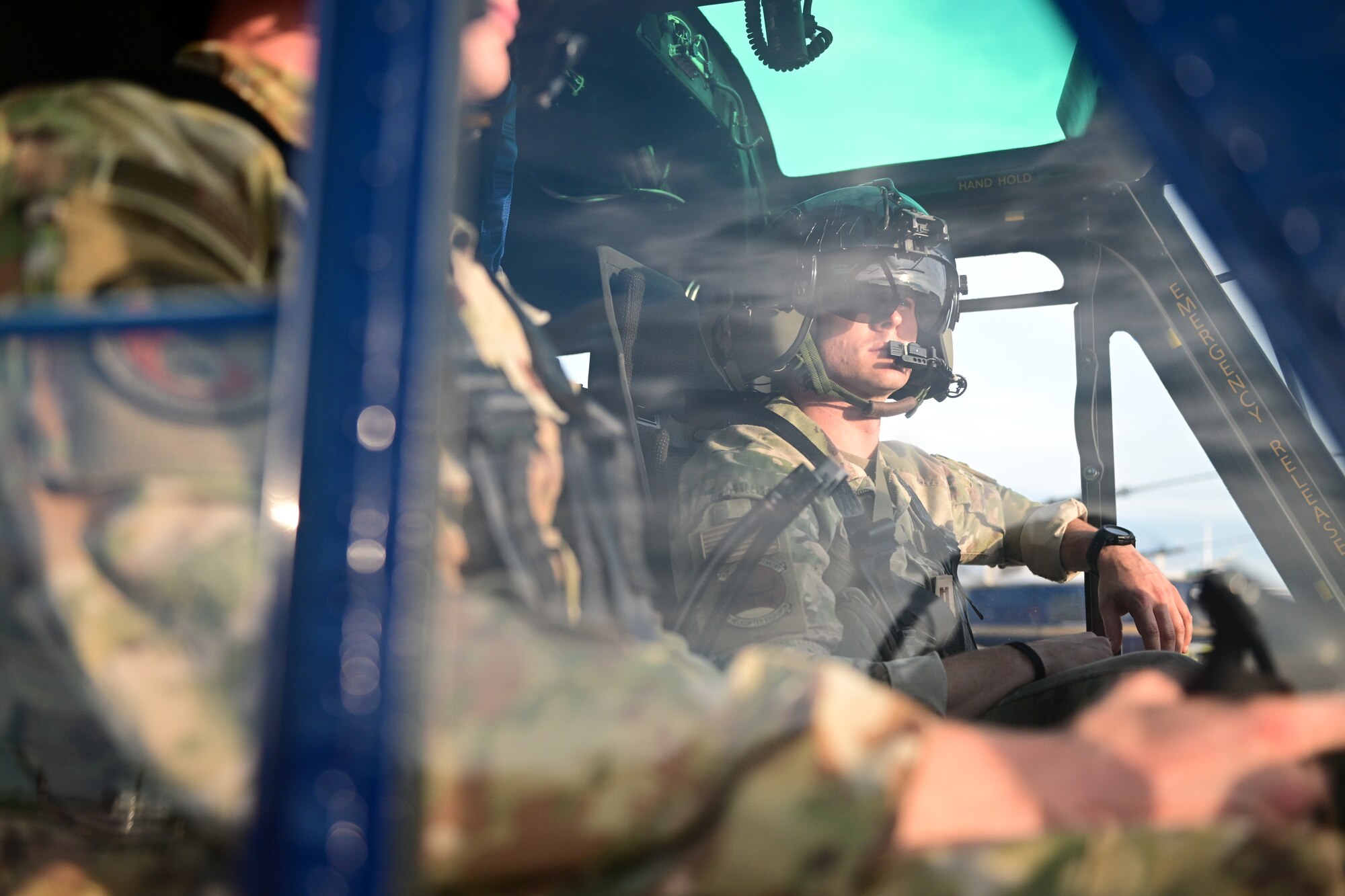 Two helicopter pilots, in military uniforms and helmets, are seen through the side window of a helicopter. One pilot is in focus, while the other is slightly blurred.