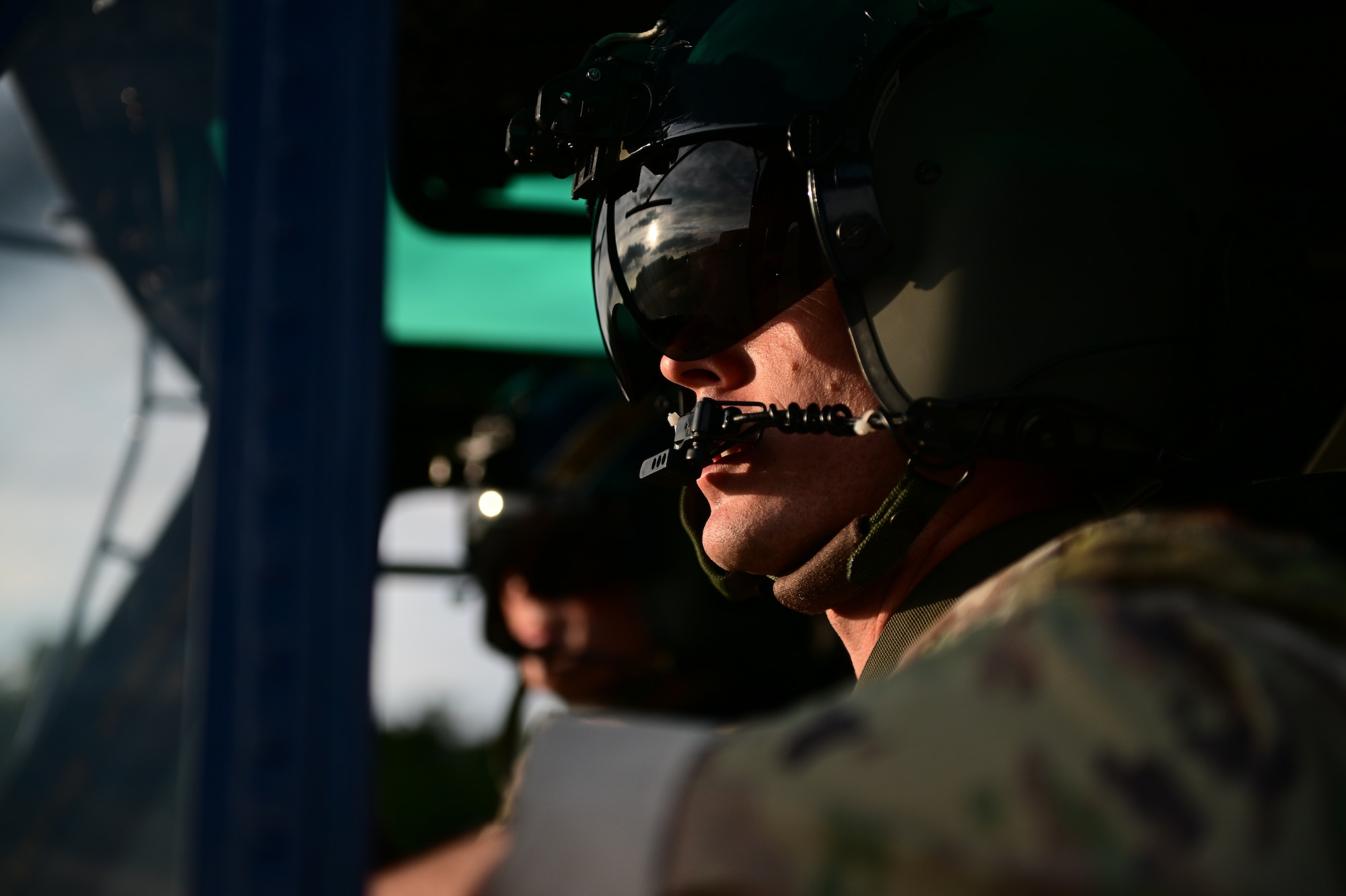 A side profile of a helicopter pilot wearing a helmet. Another pilot is visible in the background, both inside the helicopter cockpit.