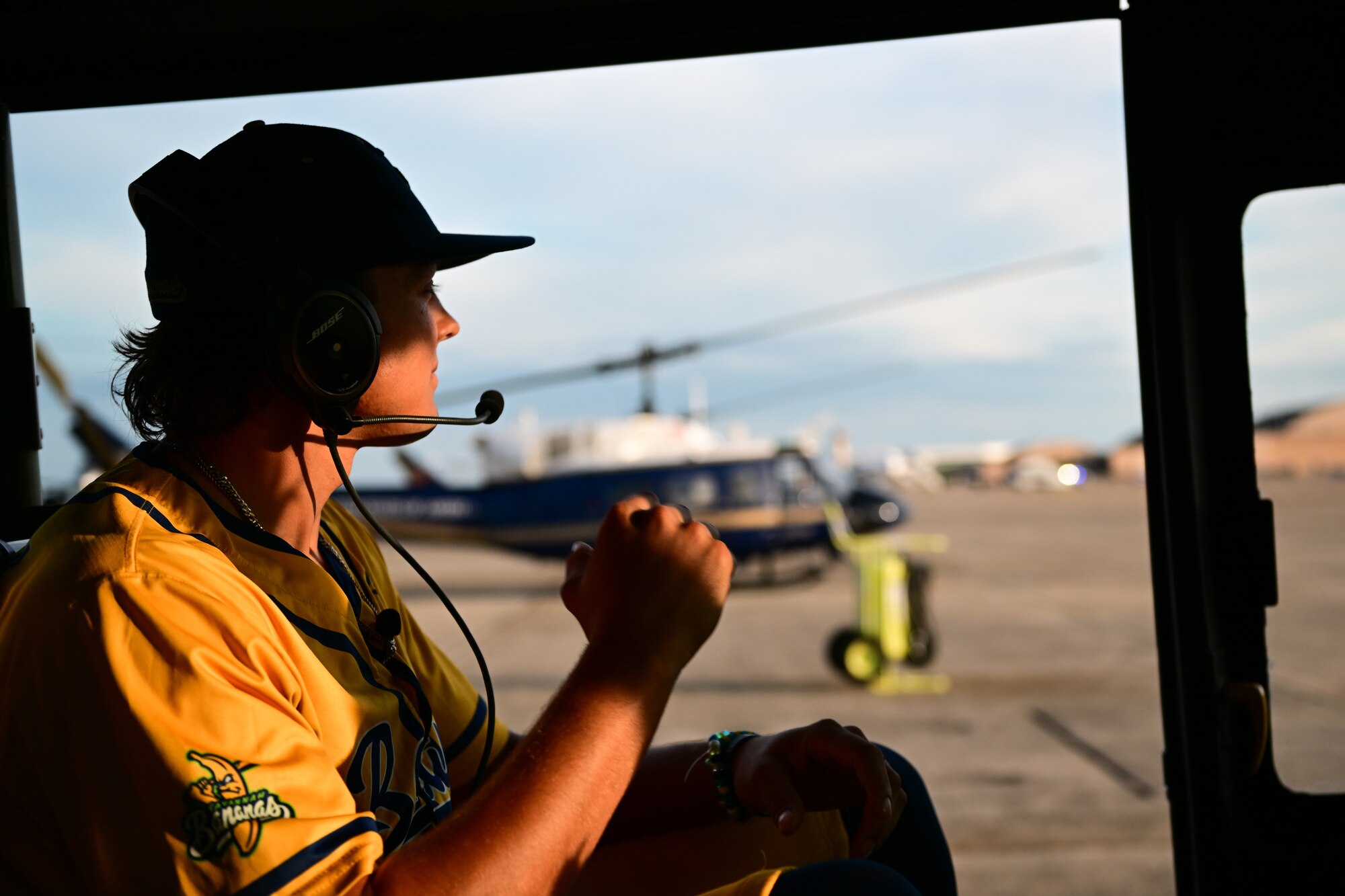 A man wearing in a yellow "Savannah Bananas" baseball jersey wearing a headset, seated inside a helicopter, with the helicopter door open.