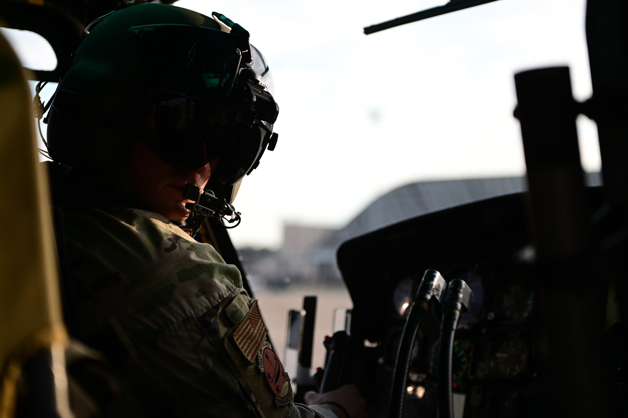 A helicopter pilot wearing a helmet looks back from the cockpit with the helicopter instruments in the foreground.