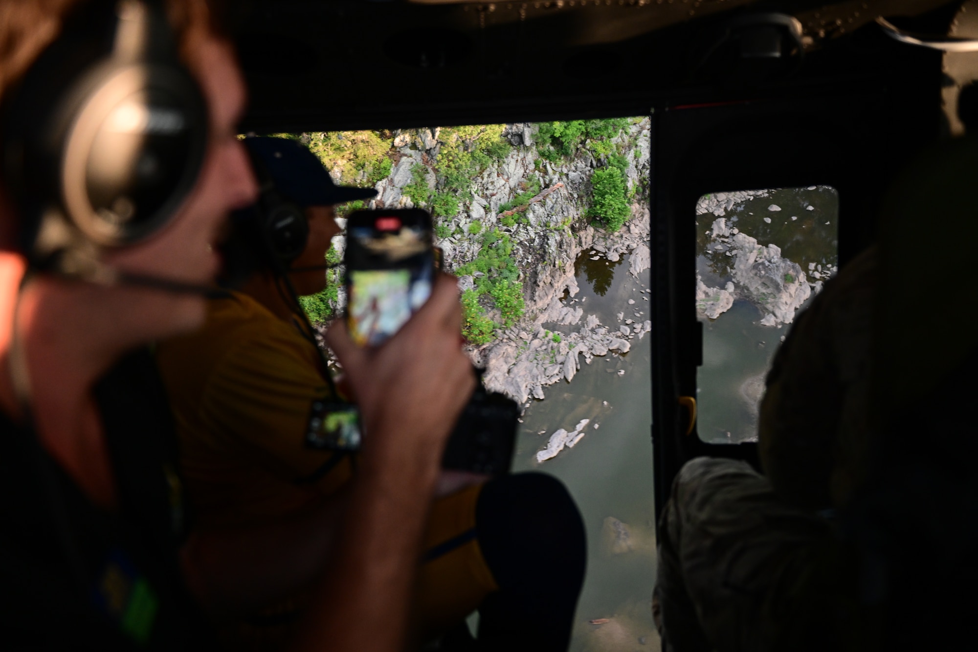 nside a helicopter, a person wearing a headset takes a photo of the landscape below, showing rocky terrain and greenery through the helicopter's open door.