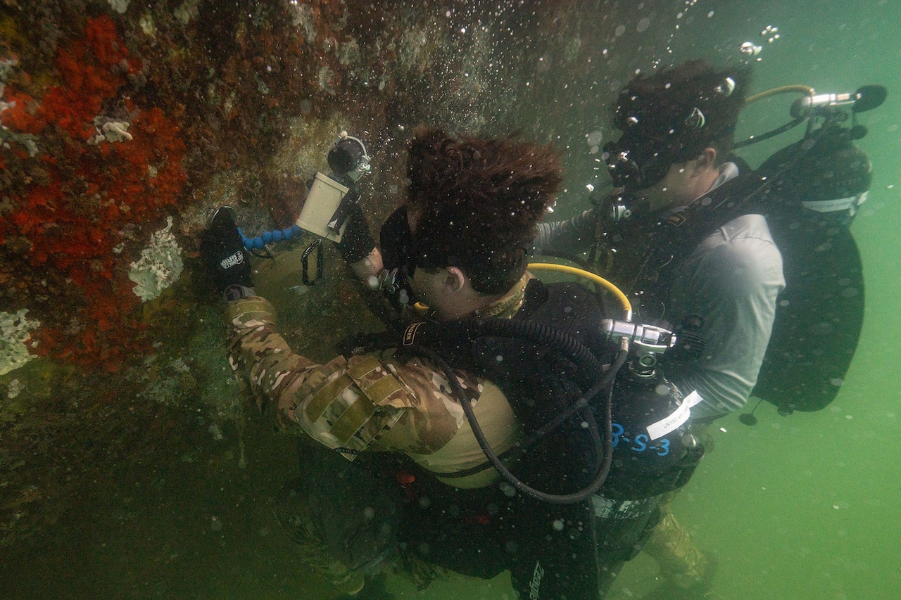 Two scuba divers with equipment touch an underwater hull covered in layers of marine sediment.