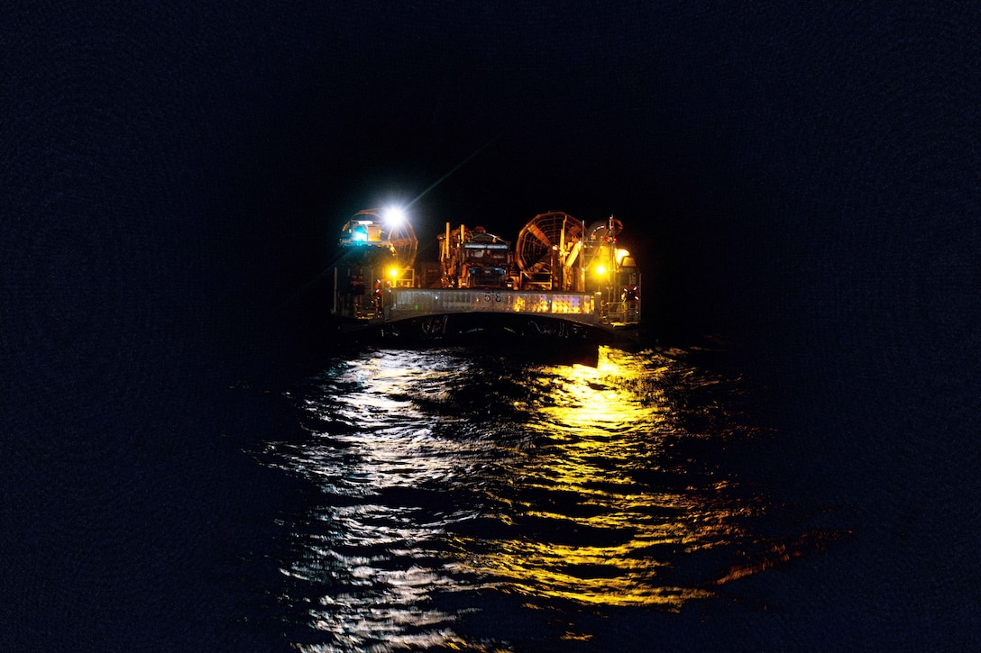 The lights from a large landing craft are seen as it moves across the water toward the camera at night.