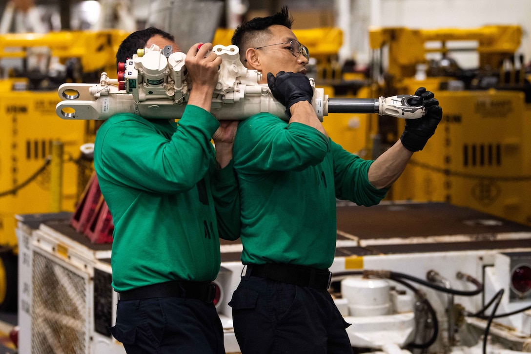 Two sailors walk close together while carrying a heavy piece of machinery on their shoulders.