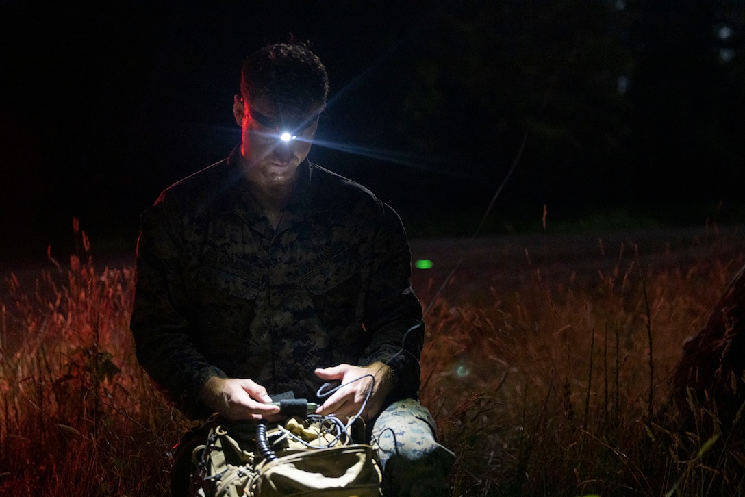 A Marine wearing a headlamp kneels in a grassy area at night to use radio equipment.