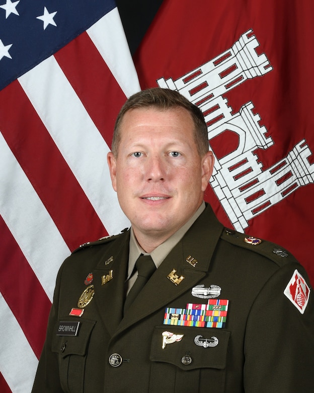 A man in an Army dress uniform smiles in front of an American flag.