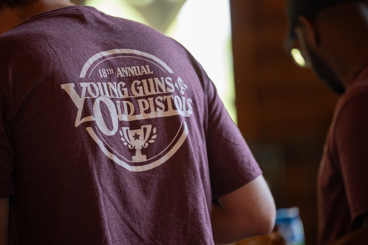 A purple t-shirt with the writing "18th Annual Young Guns vs. Old Pistols" is pictured.