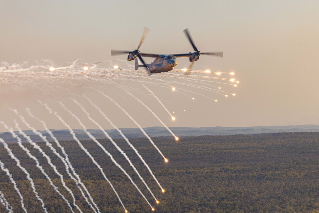 Marines in an airborne helicopter release dozens of flares over trees with mountains in the distance.