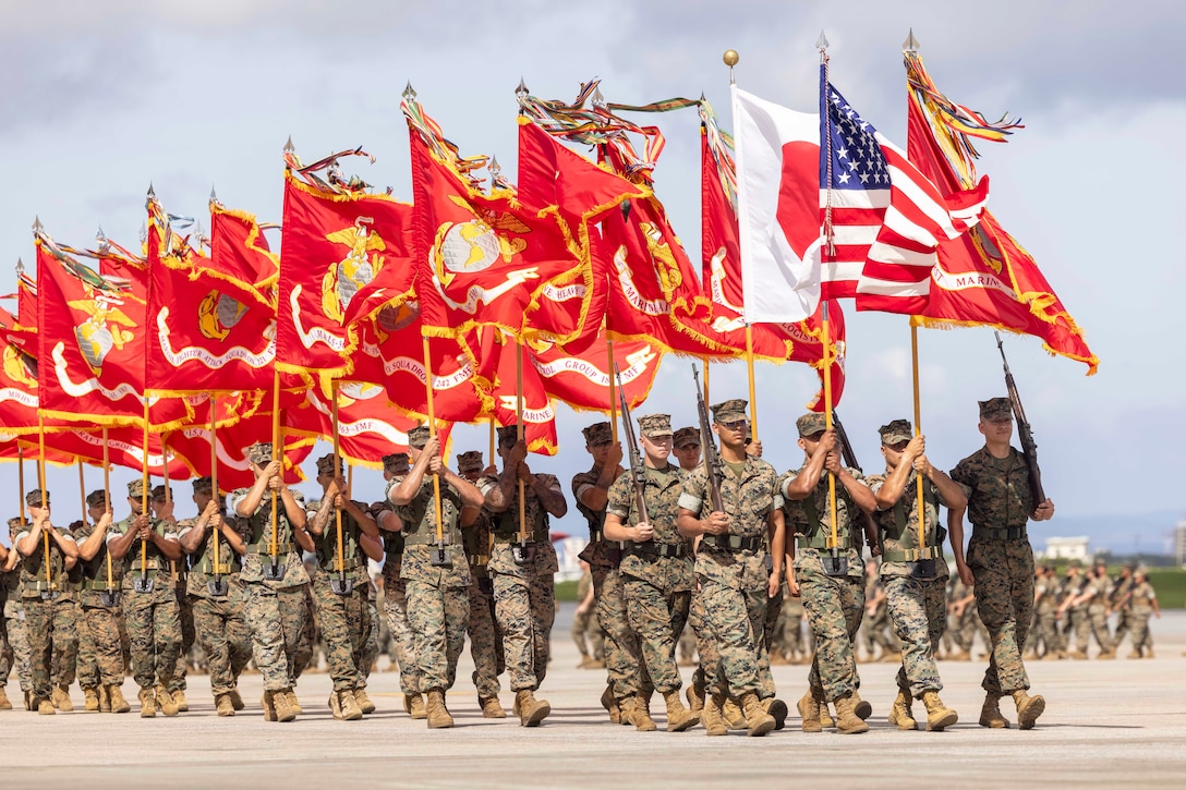 A Marine carrying an American flag leads dozens of Marines marching in formation carrying red, orange and white flags.