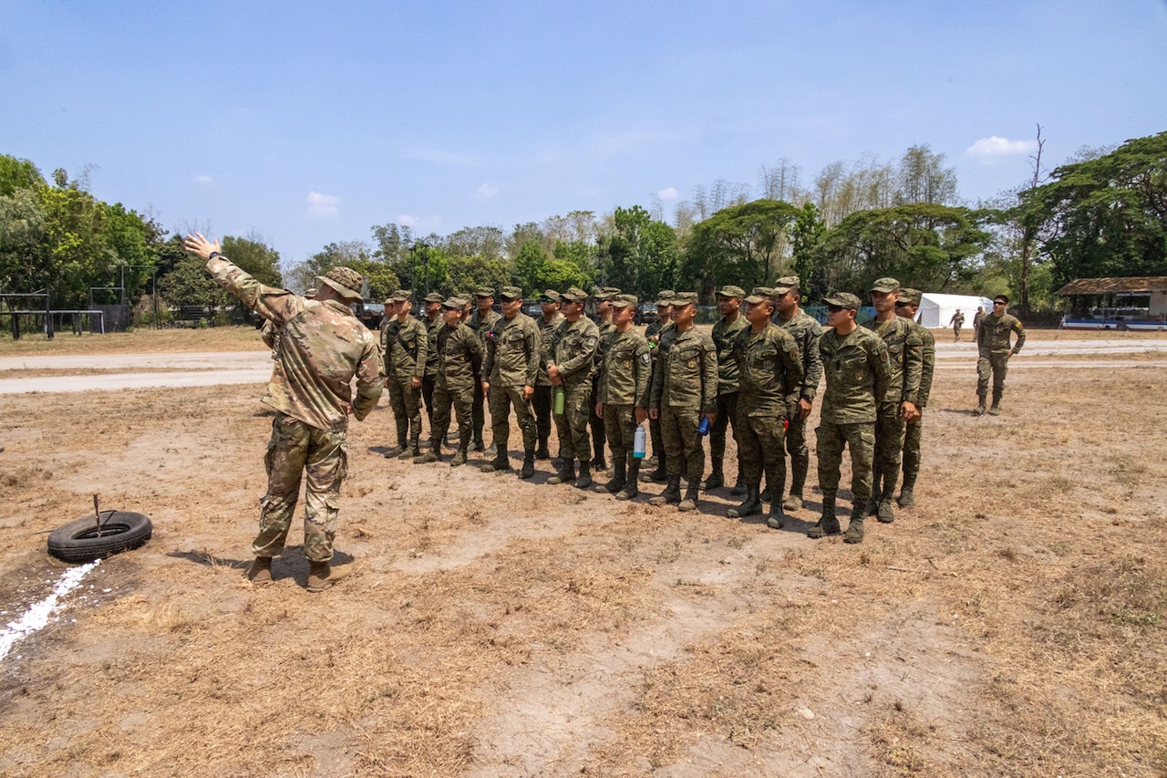 A service member holds their arm in the air as they address foreign service members standing in formation.
