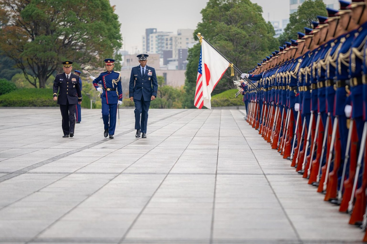 A U.S. service member in dress uniform walks between two people wearing military uniforms as an honor cordon stands at attention.