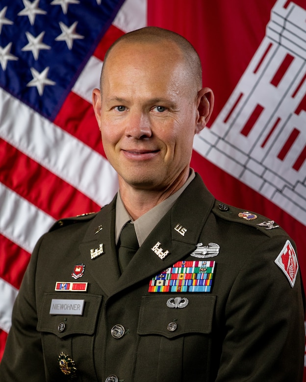 A man in a dress Army uniform smiles in front of an American flag.