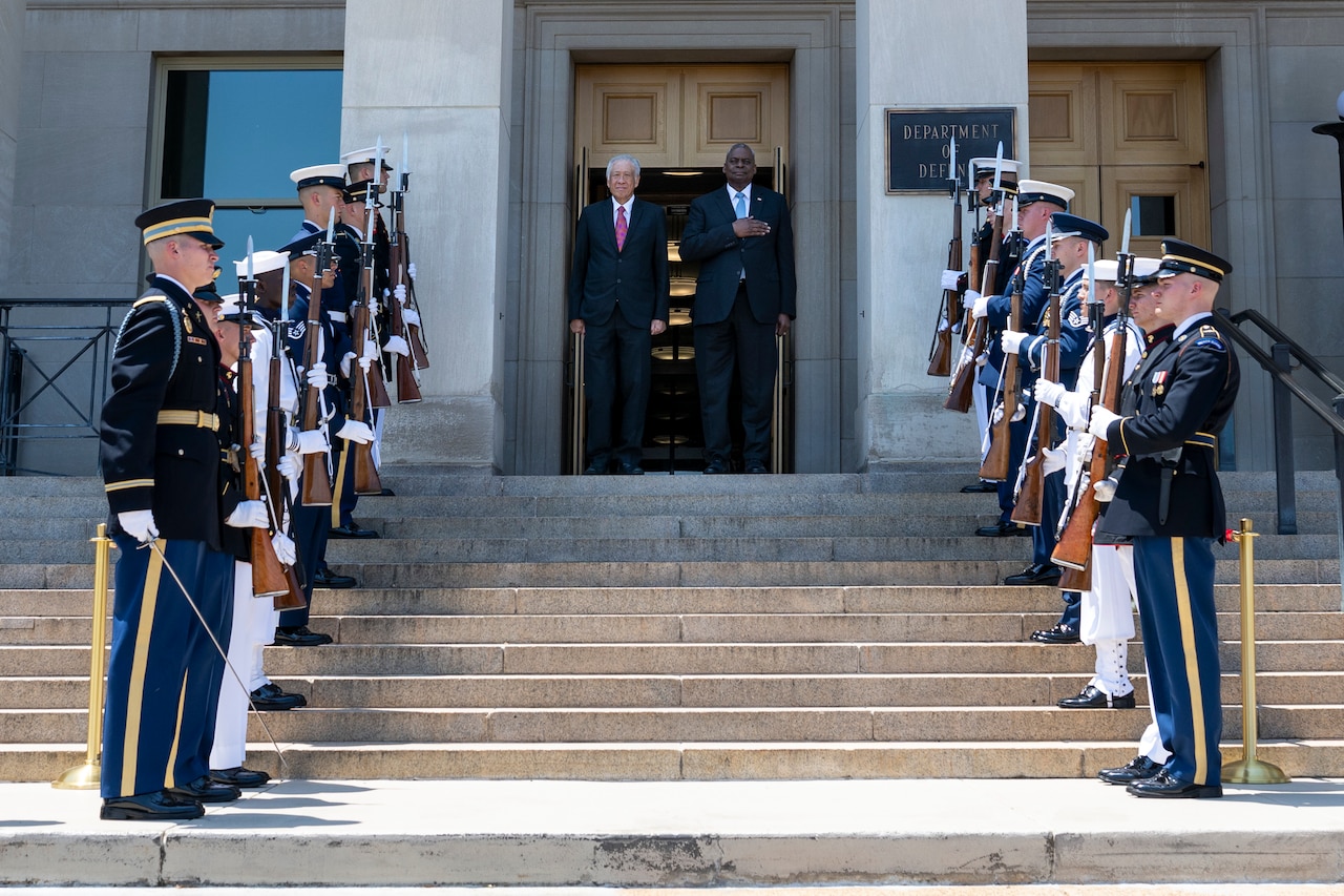 Two people stand on the steps on the Pentagon surrounded by service members in ceremonial dress.