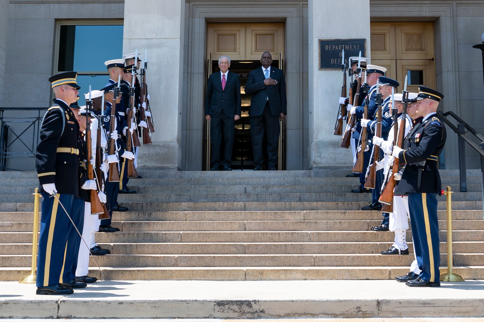 Two people stand on the steps on the Pentagon surrounded by service members in ceremonial dress.