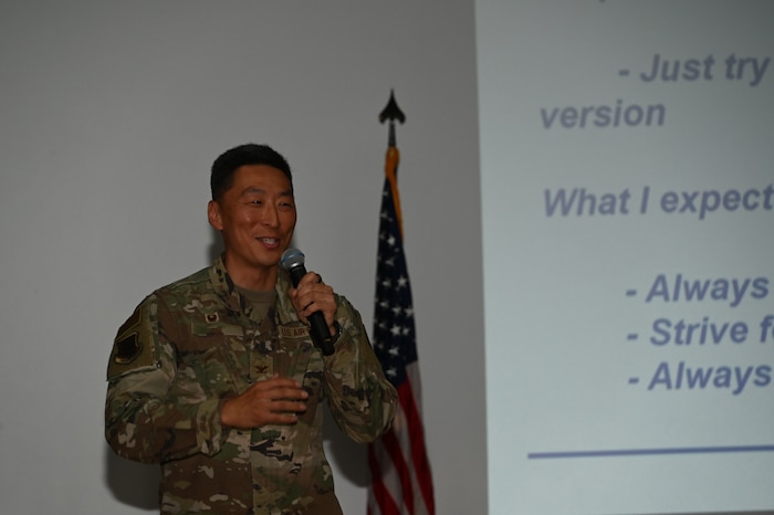 U.S. Air Force Service Member speaks into a microphone he is holding with his left hand while in front of an American flag.