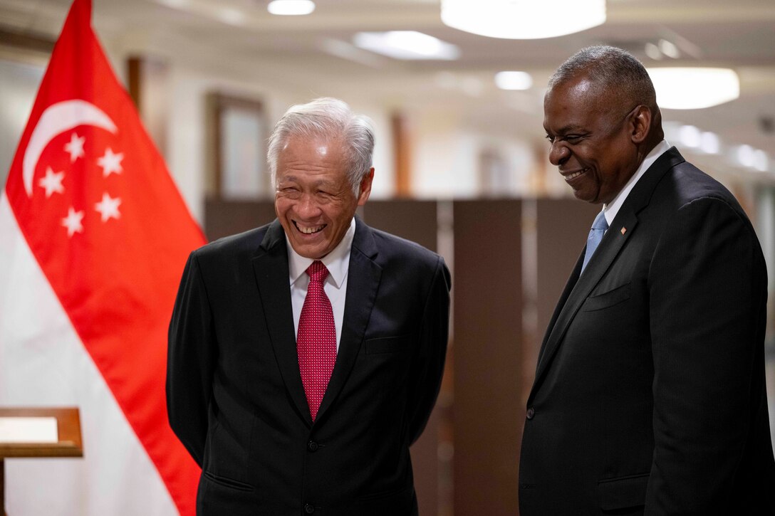 Secretary of Defense Lloyd J. Austin III laughs with his Singapore counterpart in front of the Singaporean flag.
